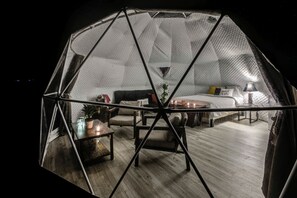 A geodesic dome room with a bed, seating area, and a small table with a plant.