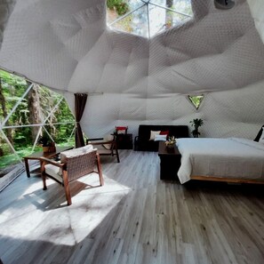 A tent-style room with a bed, a chair, a small table, and a view of the forest.