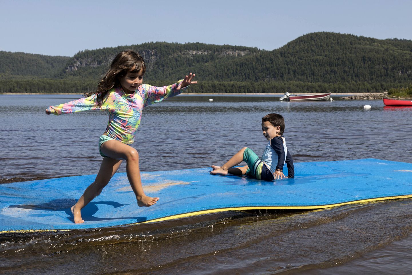 Two children on a blue mat by a lake with boats and hills in the background.