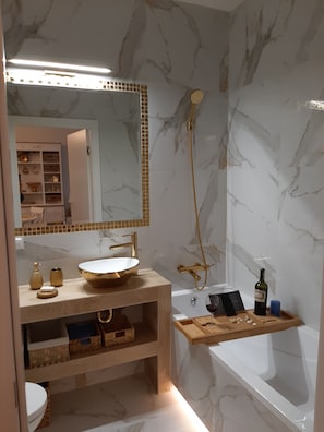 Une salle de bain avec un mur en marbre, un lavabo avec un receveur doré, une douchette à main et une étagère en bois où sont rangés les articles de toilette.