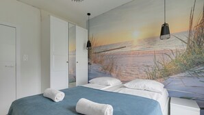 A bedroom with a bed, two bedside lamps, a white wardrobe, and a wall mural of a beach scene. - Riverfront by Grand Apartments (Gdańsk)