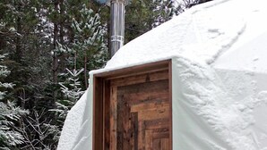 En yurt dækket af sne med en trædør, skorsten der ryger, og en vævet taske hængende på siden.