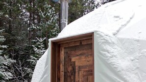 Una yurta cubierta de nieve con puerta de madera, una chimenea que emite humo y un bolso tejido colgado a un lado.