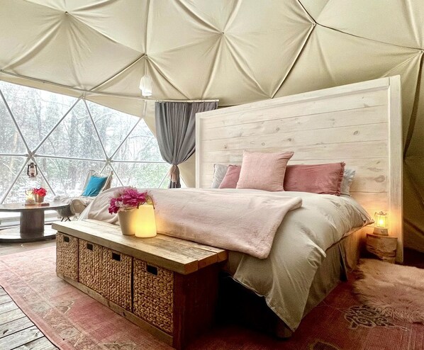 A cozy bedroom with a wooden headboard, a bed with pink bedding, a bedside table with a lamp, and a window with curtains.