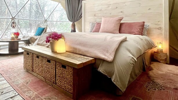 Et hyggeligt soveværelse med sengegavl i træ, seng med pink sengetøj, et natbord med lampe og et vindue med gardiner.