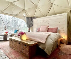 A cozy bedroom with a wooden headboard, a bed with pink bedding, a bedside table with a lamp, and a window with curtains.
