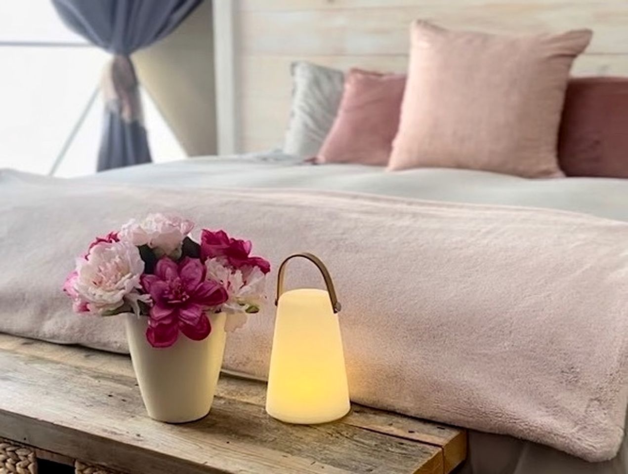 A bed with a pink blanket, a vase with flowers, and a lit lantern on a wooden table.