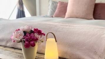 A bed with a pink blanket, a vase with flowers, and a lit lantern on a wooden table.