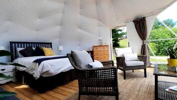 A tented accommodation with a bed, chairs, a table, and a view of greenery.
