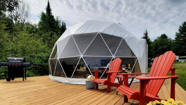 A geodesic dome tent with a wooden deck, red Adirondack chairs, a grill, and a potted plant.