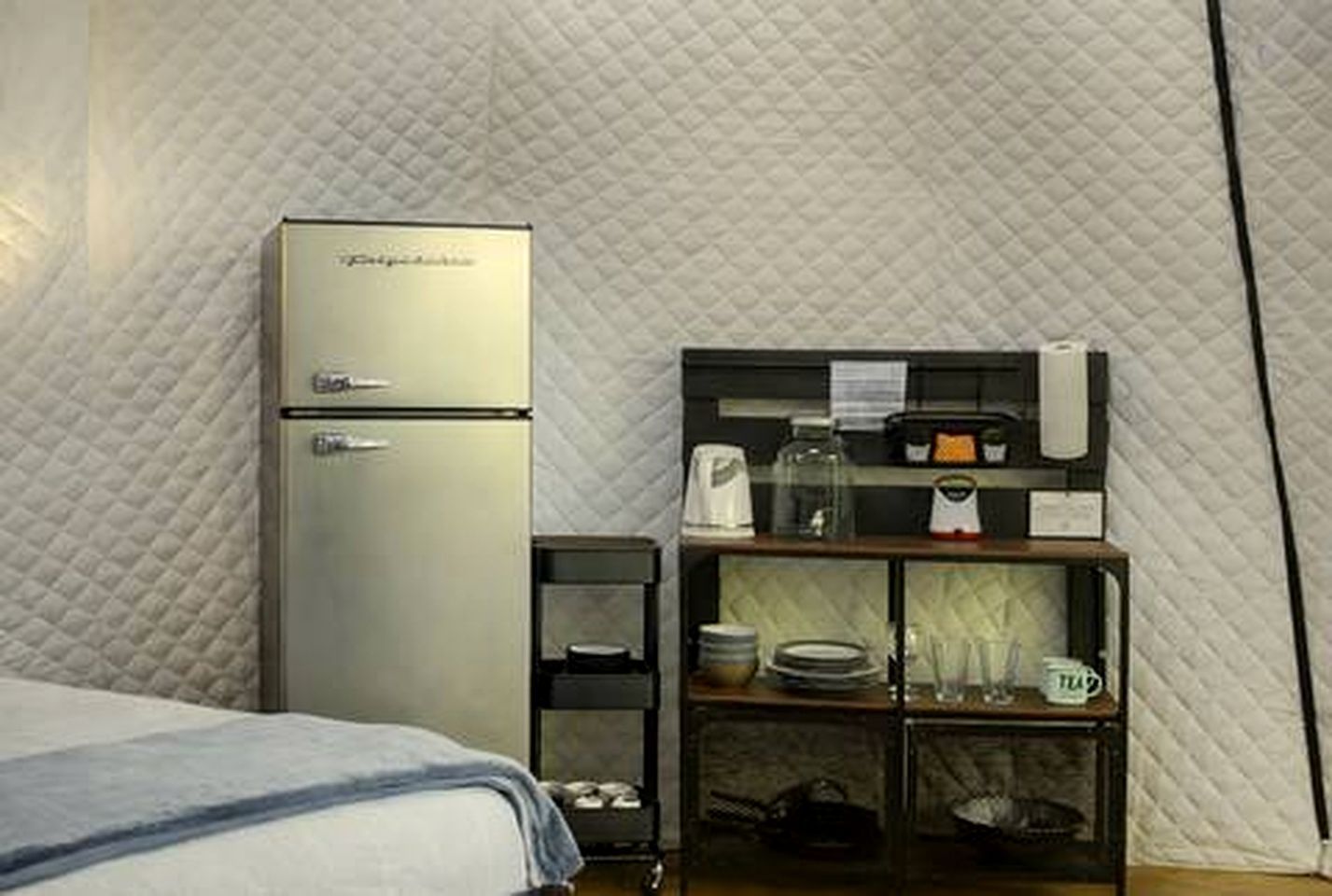 A compact room with a bed, a kitchenette area including a refrigerator, a coffee maker, and a small shelf with dishes.