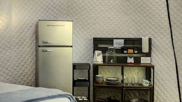 A compact room with a bed, a kitchenette area including a refrigerator, a coffee maker, and a small shelf with dishes.