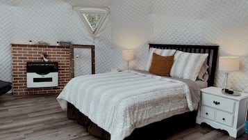 A bedroom with a quilted ceiling, a brick fireplace, a bed with a white duvet, and a nightstand with a lamp.