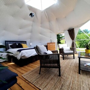 A tented accommodation with a bed, chairs, a table, and a view of greenery.