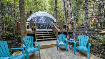 Une structure en forme de dôme au cœur d’une forêt, une terrasse en bois avec des chaises et un escalier en bois menant à une plateforme.