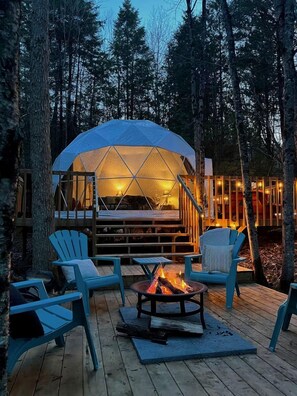 A dome-shaped tent with a bed inside, surrounded by wooden deck furniture and a fire pit.