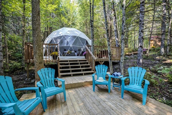 A dome structure in a forest, wooden deck with chairs, and a wooden staircase leading to a platform.