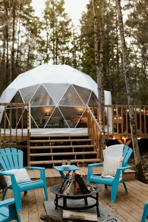 A geodesic dome structure with a wooden deck, two blue chairs, and a fire pit.