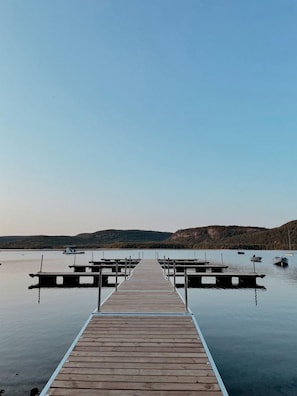 Un molo di legno si protende su acque calme, con barche ormeggiate e un paesaggio collinare sullo sfondo.