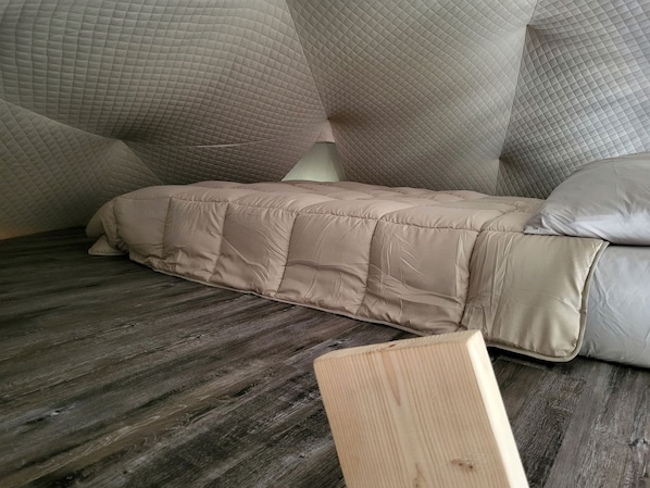 A bed with a quilted mattress and pillows, a wooden floor, and a visible wooden headboard.