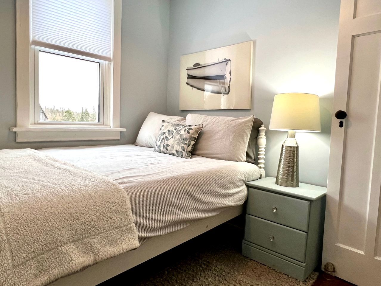 A bedroom with a bed, a nightstand with a lamp, a window with blinds, and a wall-mounted picture.