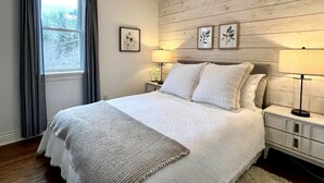 Un dormitorio con cama, dos mesitas de noche, ventilador de techo y ventana con cortinas.