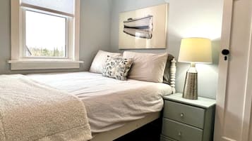 A bedroom with a bed, a nightstand with a lamp, a window with blinds, and a wall-mounted picture.