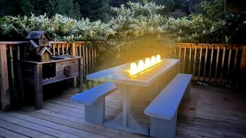 Une terrasse en bois avec une table et des bancs, entourée d’une clôture en bois et d’un feuillage dense.