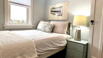 A bedroom with a bed, a nightstand with a lamp, a window with blinds, and a wall-mounted picture.