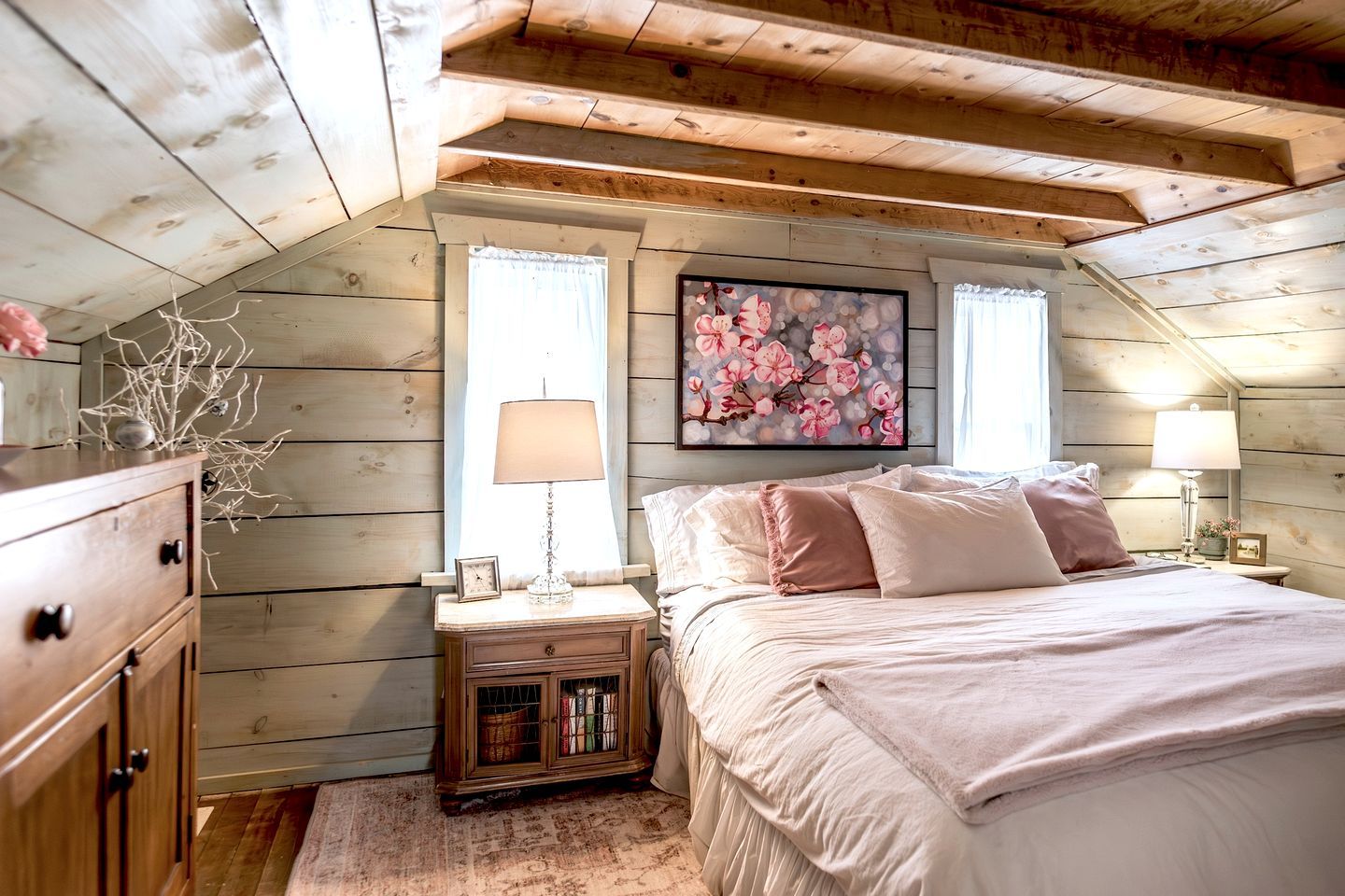 A cozy bedroom with a sloped ceiling, wooden walls, a bed with pink bedding, two bedside lamps, a nightstand with a lamp and books, and a floral painting on the wall.