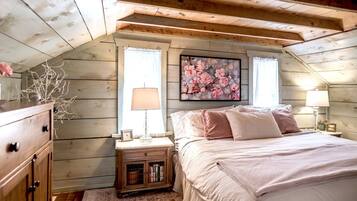 A cozy bedroom with a sloped ceiling, wooden walls, a bed with pink bedding, two bedside lamps, a nightstand with a lamp and books, and a floral painting on the wall.
