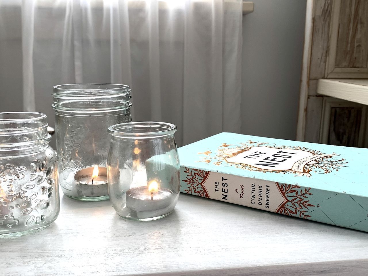A book titled 'The Nest' by Cynthia D'Aprix Sweeney, accompanied by three lit candles in glass jars on a table.