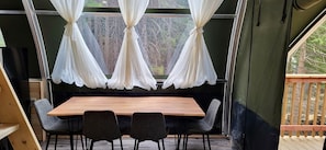 A dining area with a wooden table and four chairs, a window with white curtains, and a view of trees outside. - Unique Tented Cabin Fully Furnished and Equipped Perfect for Groups or Families in Alberta (Beaver Mines)