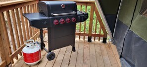 A portable grill with four burners and a gas tank on a wooden deck.