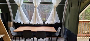 A dining area with a wooden table and four chairs, a window with white curtains, and a view of trees outside.
