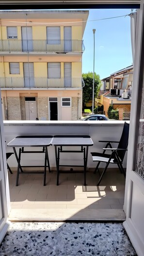En åben balkon med et bord og stole, udsigt til en gul bygning og en gade med parkerede biler.