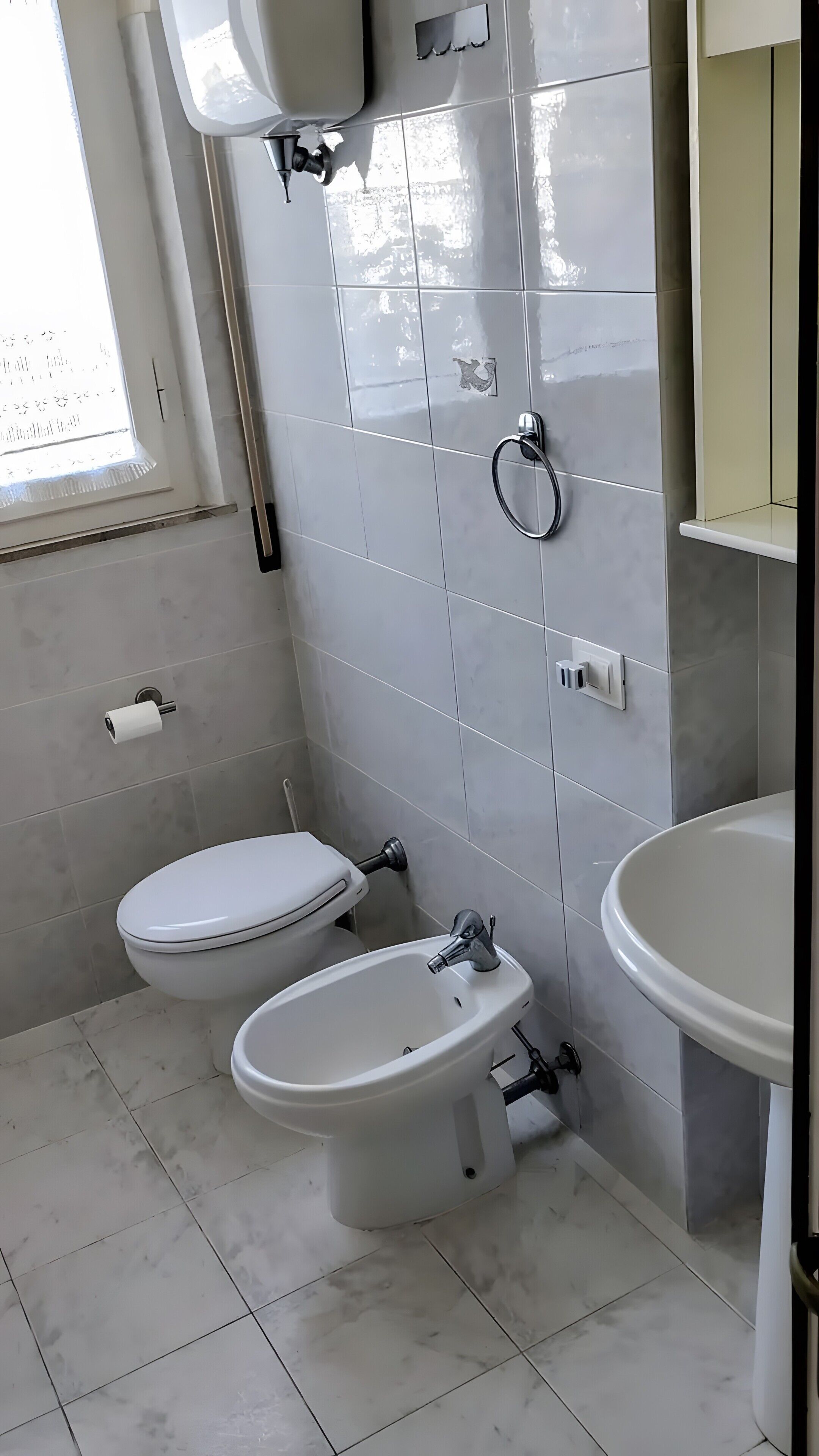Et badeværelse med toilet, bidet og vask, flisemure og et vindue.