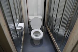 Un baño pequeño y estrecho, con un inodoro blanco, un dispensador de papel higiénico y un desagüe en el suelo.