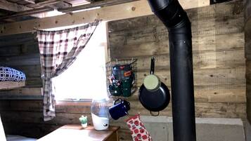 A rustic cabin interior with a wooden table, a window with plaid curtains, a hanging pot rack, and a black chimney.