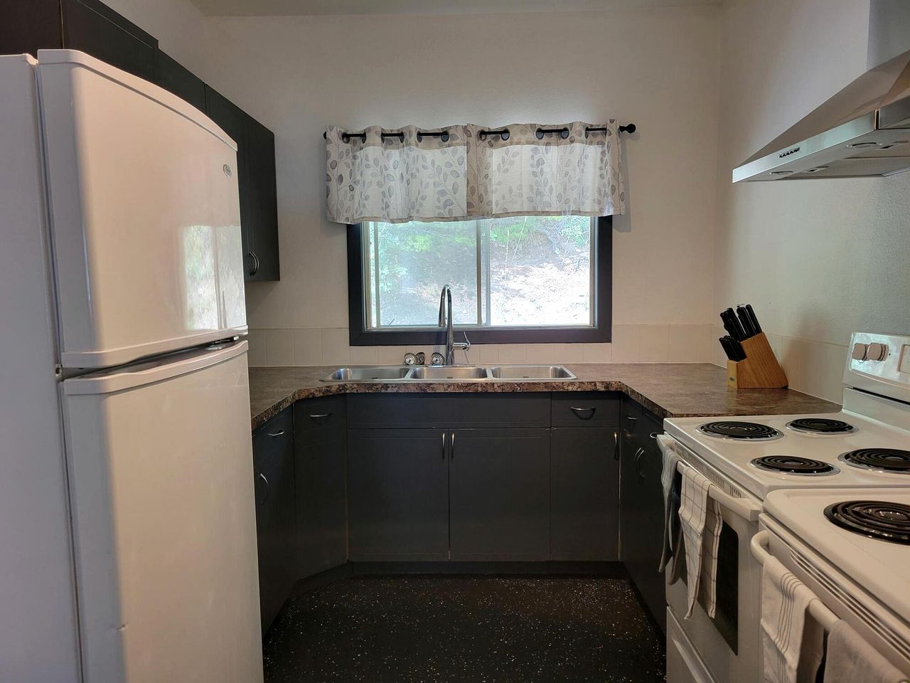 A compact kitchen with a refrigerator, stove, sink, and window with a curtain.