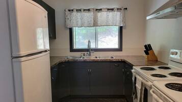 A compact kitchen with a refrigerator, stove, sink, and window with a curtain.