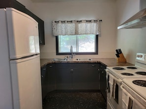 A compact kitchen with a refrigerator, stove, sink, and window with a curtain.