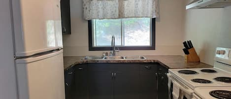 A compact kitchen with a refrigerator, stove, sink, and window with a curtain.