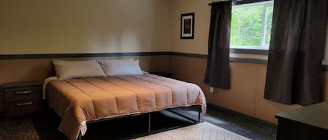 Een slaapkamer met een bed, een nachtkastje, een dressoir, een raam met gordijnen, en een lijst aan de muur.