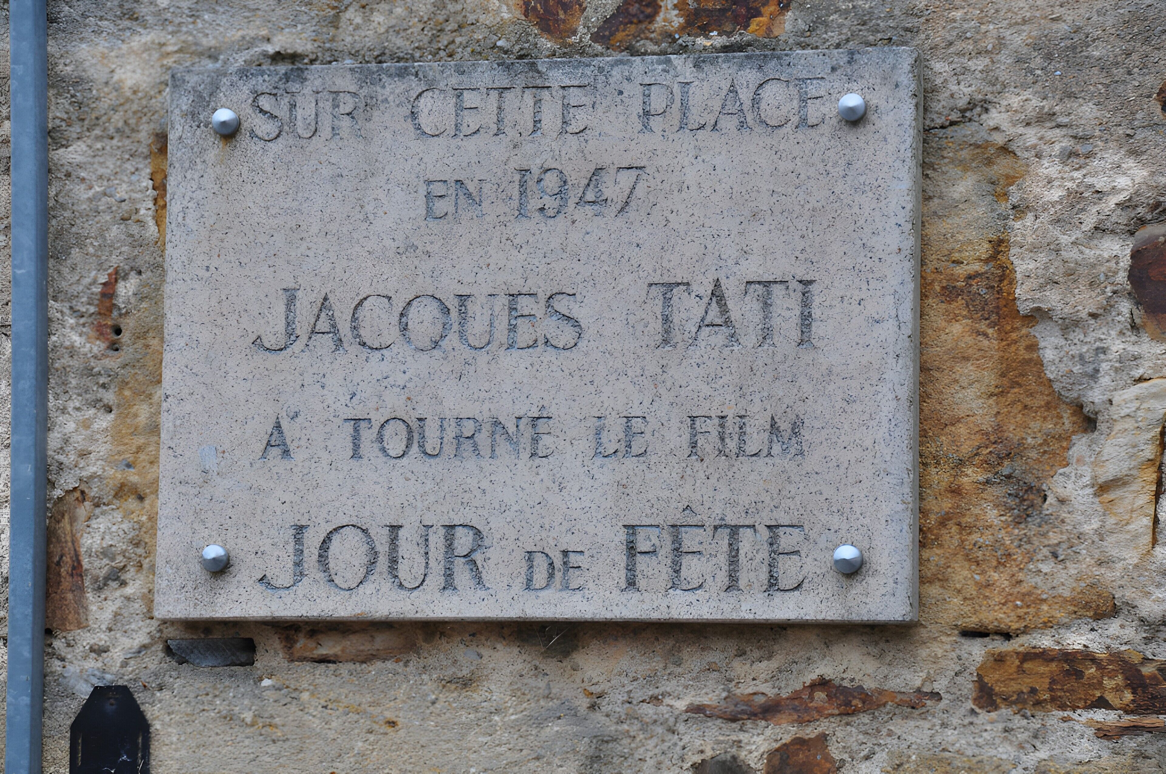 Une plaque de pierre usée par les intempéries, portant des inscriptions, fixée à un mur qui présente des signes d’usure et de rouille.