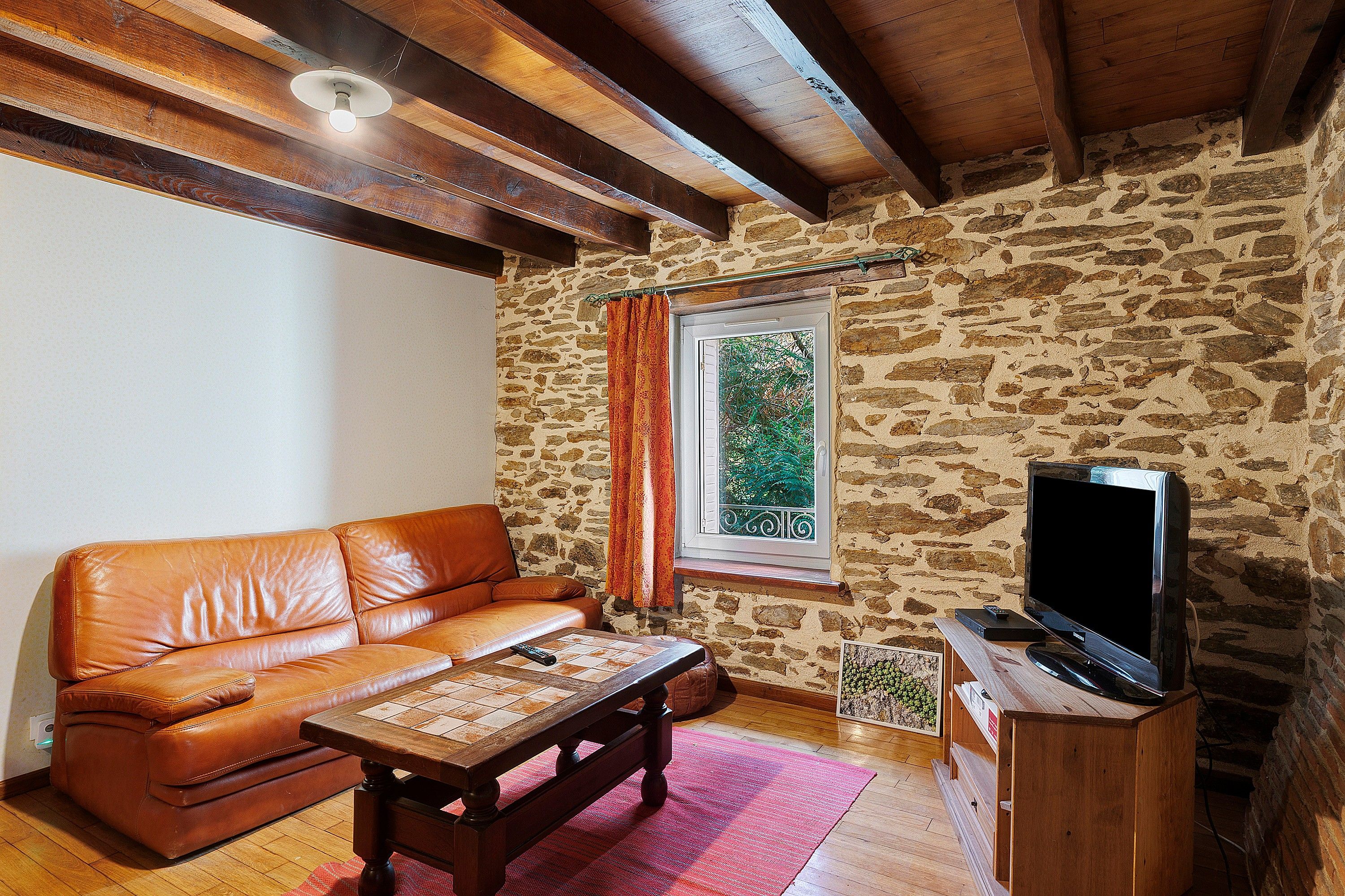 A cozy living room with a leather sofa, a wooden coffee table, a television on a wooden stand, and a stone wall.