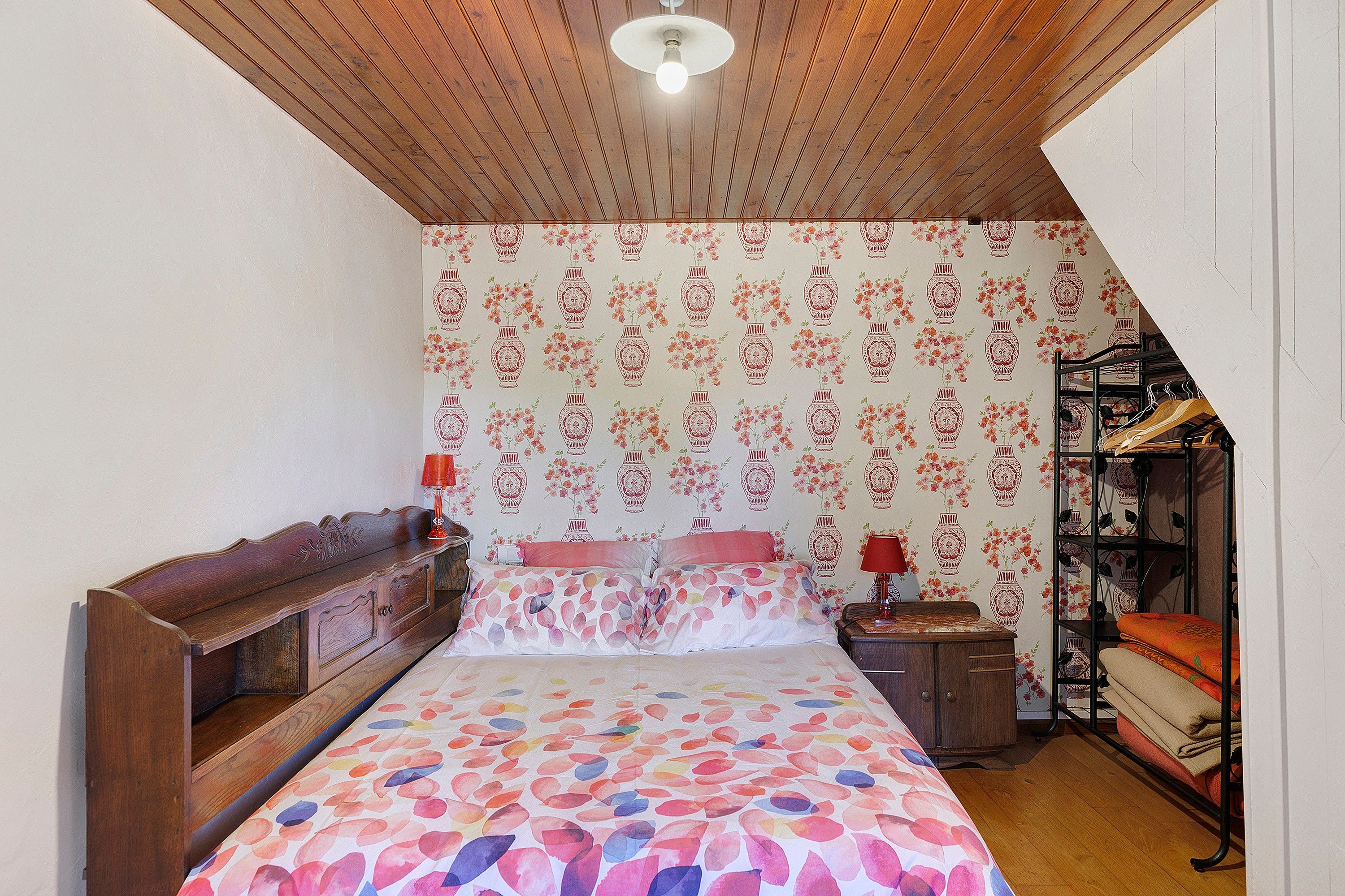 A bedroom with a wooden ceiling, a bed with floral bedding, a nightstand with a lamp, and a wardrobe.