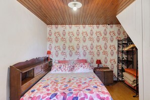 Une chambre avec un plafond en bois, un lit avec une literie à motifs floraux, une table de chevet avec une lampe et une armoire.