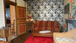 A cozy living room with a wooden floor, a red rug, a striped sofa, a wicker chair, and a fireplace. - Holiday Home “La Charmille” with Private Terrace and Garden (Nouzerolles)