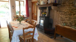 A dining area with a table set for four, chairs, a vase with flowers, a stone wall, and a wood-burning stove. - Holiday Home “La Charmille” with Private Terrace and Garden (Nouzerolles)